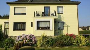 Ein zweistöckiges Haus mit einem Balkon, einem Fenster mit Vorhang und einem Blumenbeet im Vorgarten.