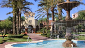 A fountain with water jets, surrounded by palm trees and a building with arched windows in the background.