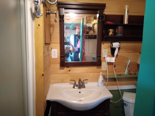 A bathroom with a wooden wall, a white sink, a mirror, a hairdryer, and a shelf with toiletries. - Lakefront Cabin on beautiful Unity Pond in Burnham, Maine (Burnham)