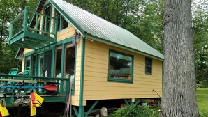 A yellow wooden cabin with a green roof and a balcony, surrounded by greenery.
