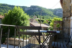 Un balcon doté d’une balustrade en métal, offrant une vue sur un village aux bâtiments traditionnels, avec une chaîne de montagnes en arrière-plan.