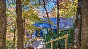 A cabin with a wooden deck and stairs leading to a blue-roofed structure surrounded by trees.