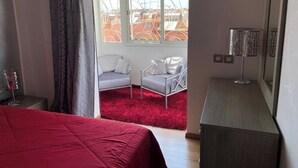 A bedroom with a red bedspread, a white chair by the window, and a lamp on a wooden nightstand. - Beautiful, luxurious and very quiet apartment. (Marrakech)