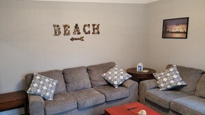A living room with two grey sofas, patterned cushions, a wooden coffee table, and a wall with the word "BEACH" made of stones.
