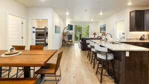 A modern kitchen with a central island, a dining area with wooden tables, and a view of the outdoors.