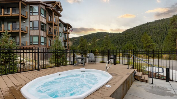 A wooden deck with a hot tub, a fenced area, and a view of a mountain and forest.