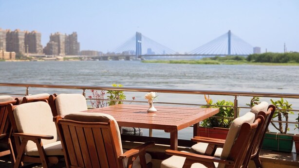 A wooden deck with chairs and a table, overlooking a river with a bridge in the background. - Nile Transport Hotel (Shubra al Khaymah)