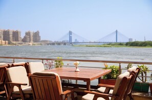 A wooden deck with chairs and a table, overlooking a river with a bridge in the background. - Nile Transport Hotel (Shubra al Khaymah)