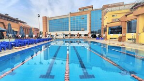 A swimming pool with blue lane dividers, surrounded by blue chairs and umbrellas, with buildings in the background. - Nile Transport Hotel (Shubra al Khaymah)