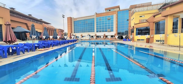 A swimming pool with blue lane dividers, surrounded by blue chairs and umbrellas, with buildings in the background.