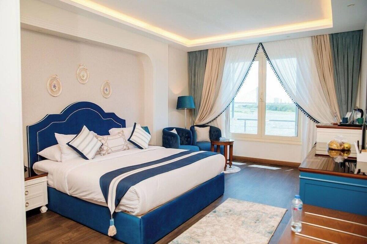 A bedroom with a large bed, a nightstand, a chair, a lamp, and a view of the outdoors.