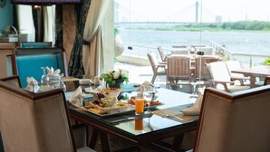 A dining area with a table set for breakfast, overlooking a body of water with a bridge in the background. - Nile Transport Hotel (Shubra al Khaymah)