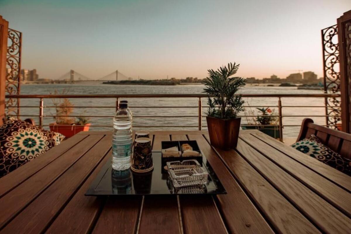 A wooden deck with a table set for two, overlooking a river with a bridge in the distance.