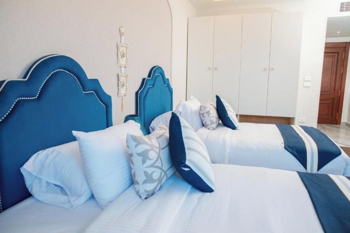 A hotel room with two beds, each with a blue headboard and white bedding. There is a wardrobe with a white door and a wooden door in the background.