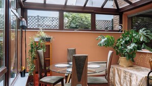 A conservatory with a glass table and chairs, surrounded by potted plants and a view of greenery.