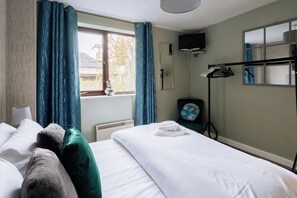 Une chambre d’hôtel avec un lit, une fenêtre avec des rideaux, un radiateur, un miroir, une télévision et un porte-vêtements.