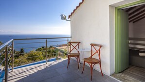 A balcony with two wooden chairs and a small table, overlooking a scenic view of the sea and hills.