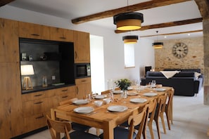 A dining area with a wooden table set for six, surrounded by wooden chairs. A stone wall with a clock is visible in the background.