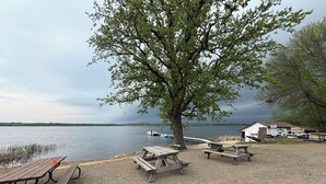 Un área de picnic junto al lago, con bancos y mesas de madera, un árbol grande y un edificio pequeño al fondo.