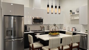 Cocina moderna con electrodomésticos de acero inoxidable, una isla central con asientos, iluminación colgante y armarios blancos.