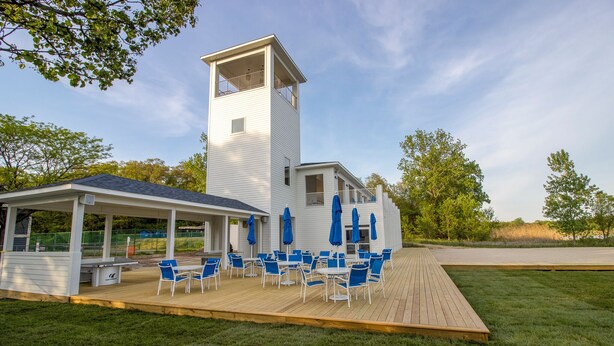 Un bâtiment blanc surmonté d’une tour, un patio couvert avec des chaises et des parasols bleus, et une terrasse en gazon.