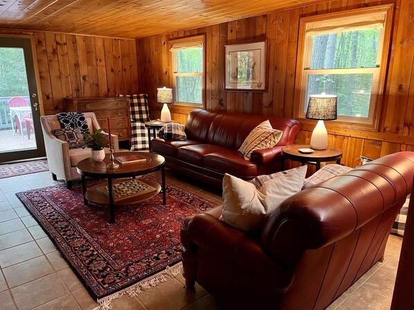 Interior acolhedor de uma cabana com um sofá de couro, uma mesa pequena e uma cadeira. Há uma janela com vista para árvores.