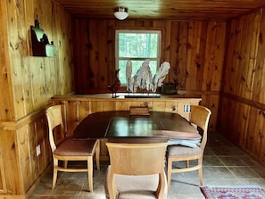 Interior de madeira com uma mesa de jantar, quatro cadeiras, uma janela com vista para árvores e um arranjo decorativo de plantas.