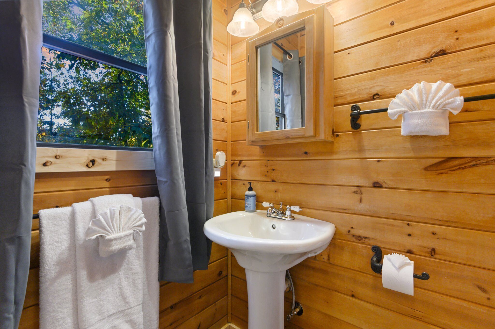 A wooden cabin bathroom with a sink, mirror, towel rack, and window with curtains.