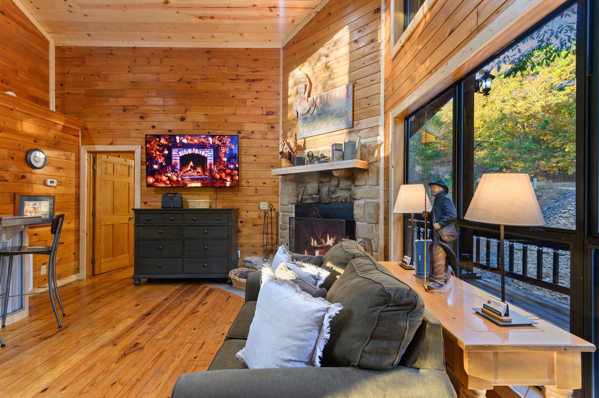 A cozy cabin interior with a stone fireplace, a television, wooden walls, and a large window with a view of trees.