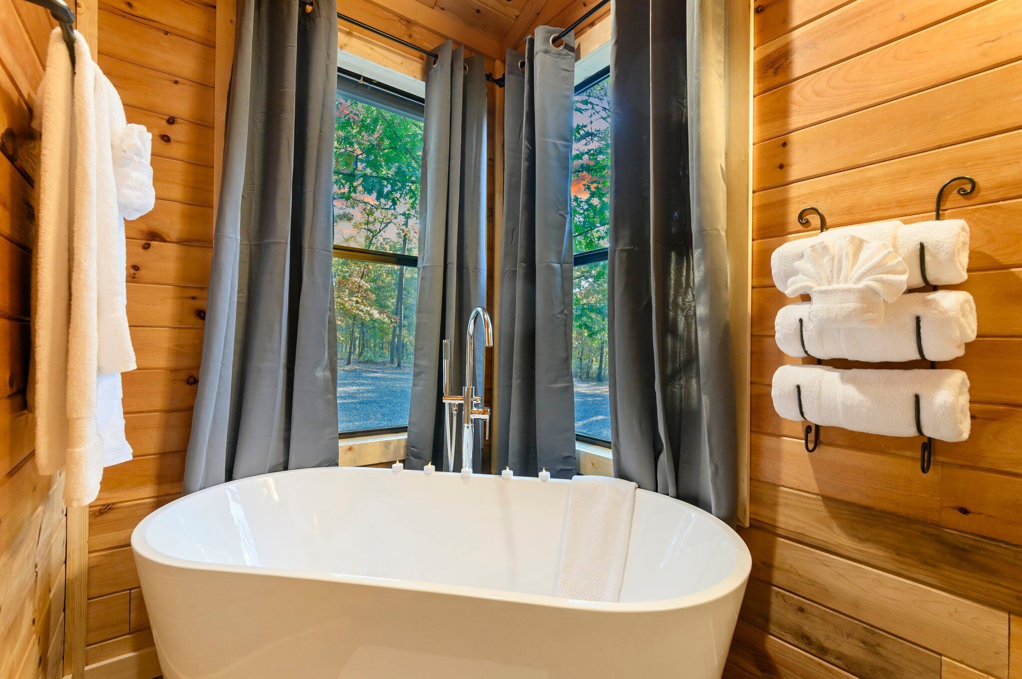A wooden cabin bathroom with a freestanding bathtub, grey curtains, and towels on hooks.