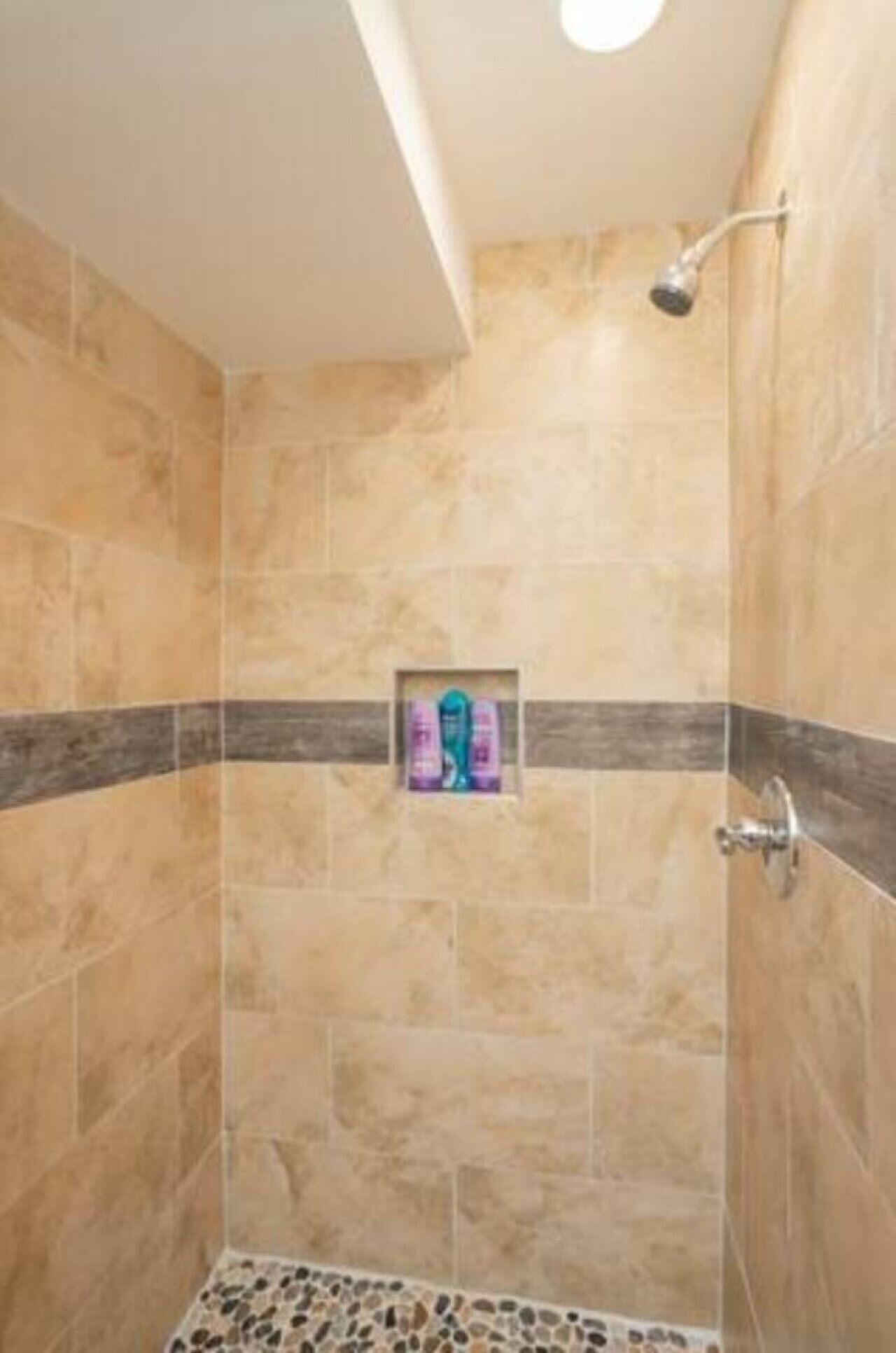 A shower with beige tiles, a dark strip, and a small shelf with toiletries.