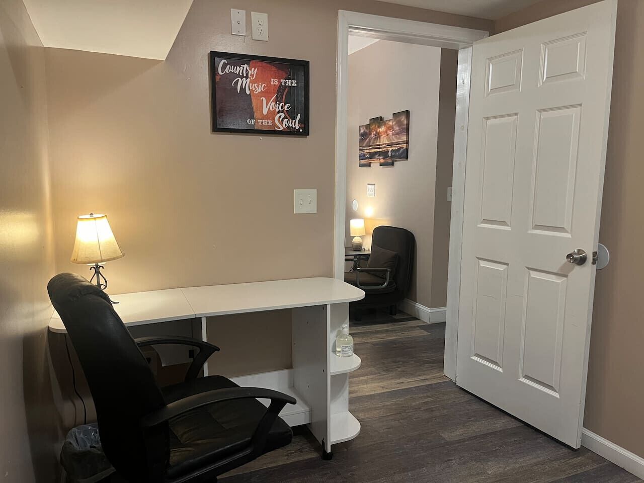 A room with a white desk, a black office chair, a lamp, and a door leading to another room. There is a poster on the wall and a small table with a chair in the background.