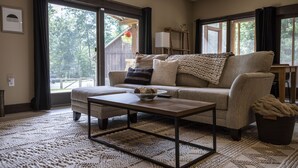 Una sala de estar acogedora con sofá, reposapiés y mesa de centro, ventanas amplias con vista a árboles, y suelo de madera.