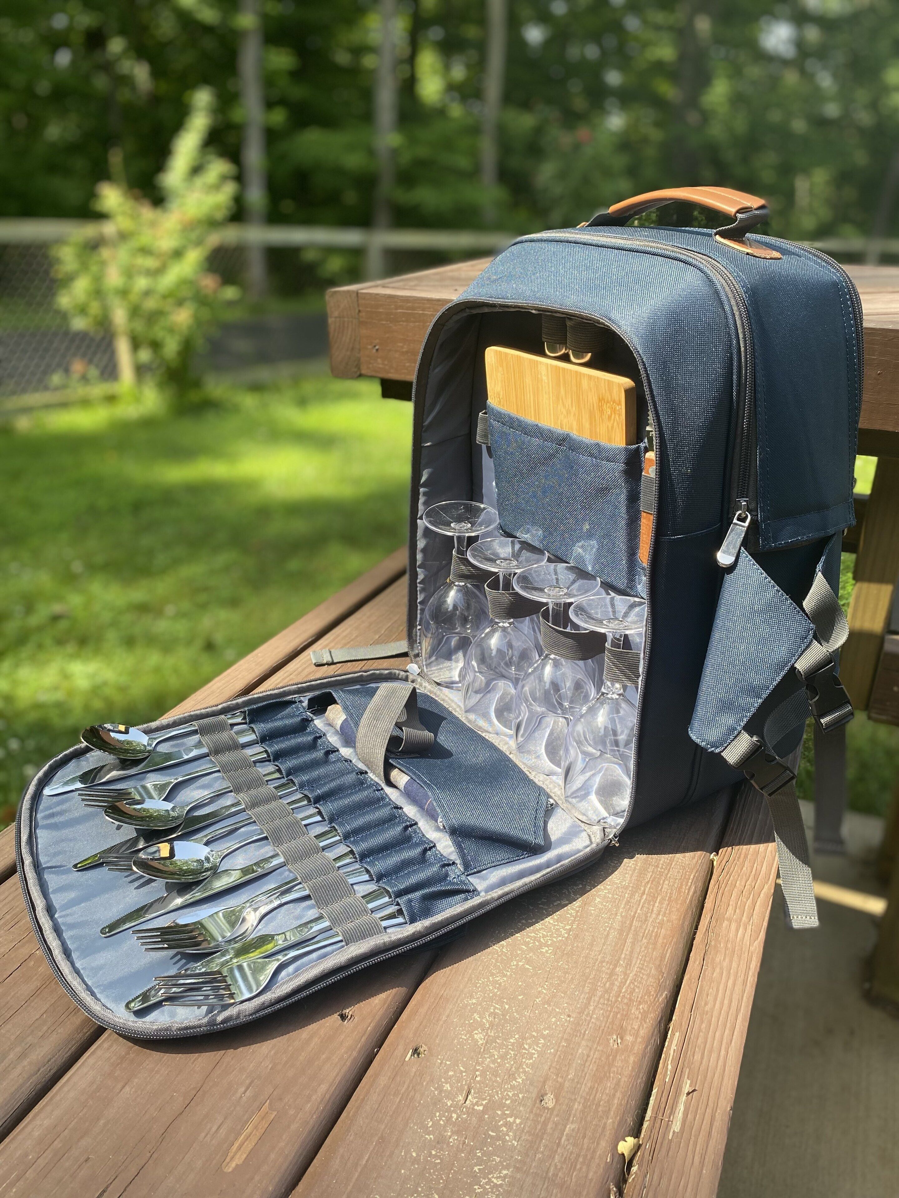 A picnic table with a backpack containing a portable grill, wine glasses, and cutlery.