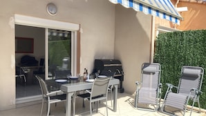 An outdoor patio with a table and chairs, a grill, and a covered area with a striped awning. - Superbe Pavillon Tout Confort Proche de la mer (Narbonne)