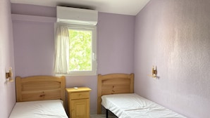 A room with two single beds, a small wooden nightstand, a window with a curtain, and an air conditioning unit. - Superbe Pavillon Tout Confort Proche de la mer (Narbonne)