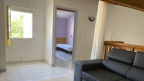 A room with a grey sofa, a white table, and a bed in the adjacent room. - Superbe Pavillon Tout Confort Proche de la mer (Narbonne)