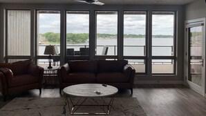 A spacious room with a large window overlooking a body of water, a ceiling fan, and a round coffee table. - Direct Lakeside View, 5 bed, 2.5 bath Condo at Bridges Bay (Arnolds Park)