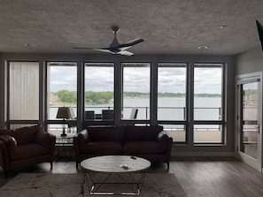 A spacious room with a large window overlooking a body of water, a ceiling fan, and a round coffee table.