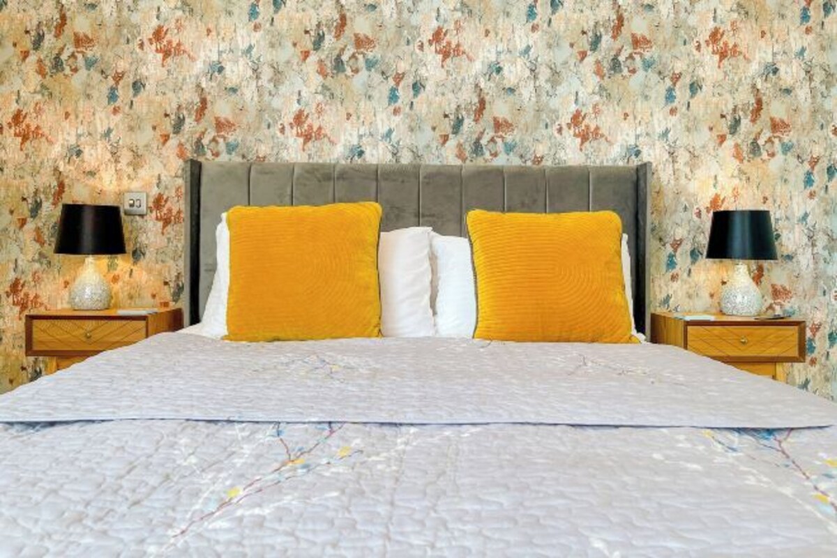 A bed with a grey upholstered headboard, two yellow pillows, and a white pillow. There are two bedside tables with lamps and decorative vases.
