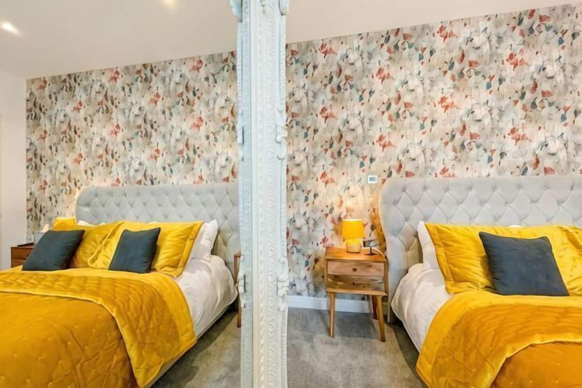 A room with two beds, floral wallpaper, a mirror, and a nightstand with a lamp.