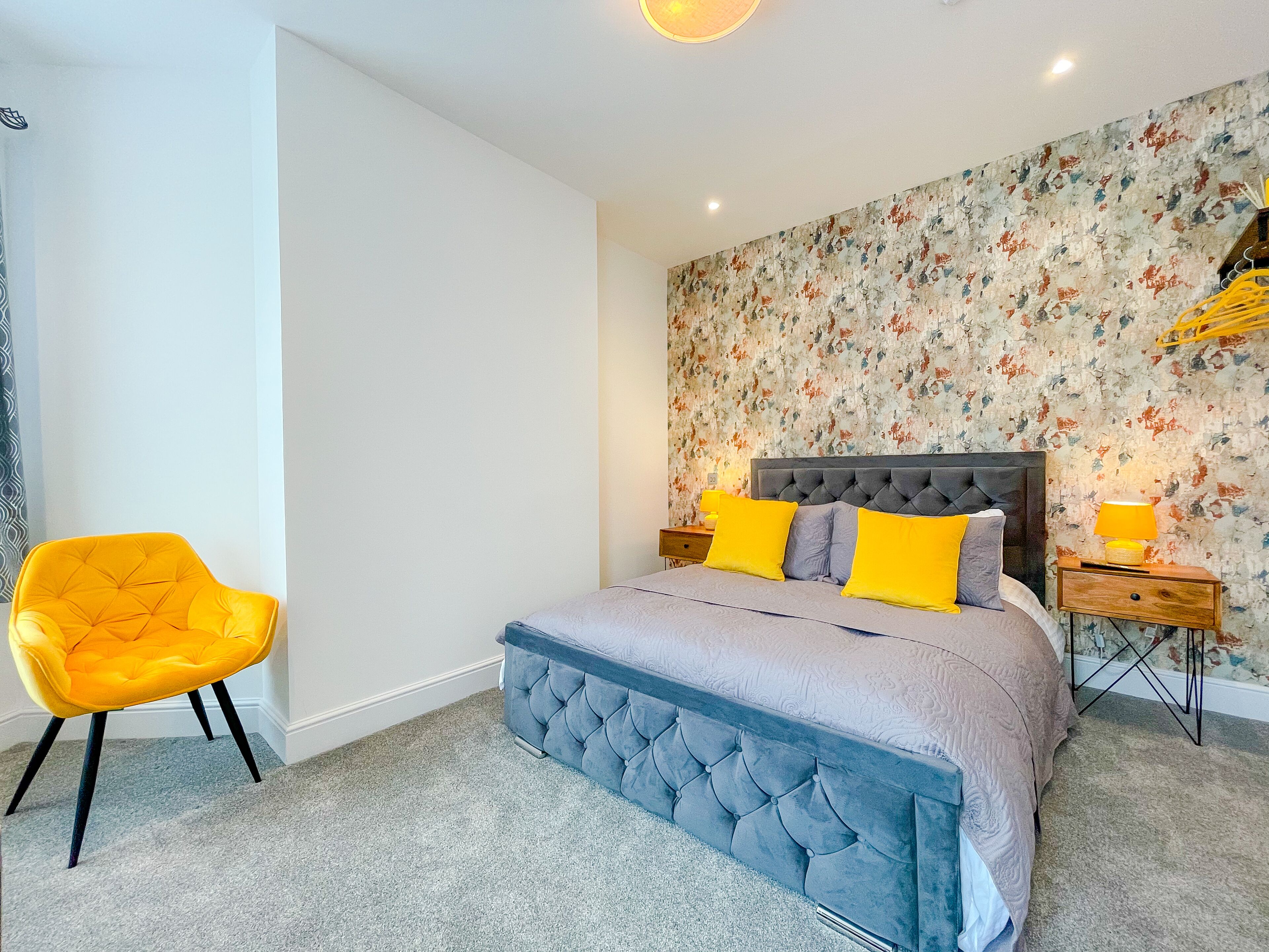 A bedroom with a tufted headboard bed, two bedside tables with lamps, a bright yellow chair, and a floral-patterned wall.