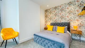 A bedroom with a tufted headboard bed, two bedside tables with lamps, a bright yellow chair, and a floral-patterned wall.