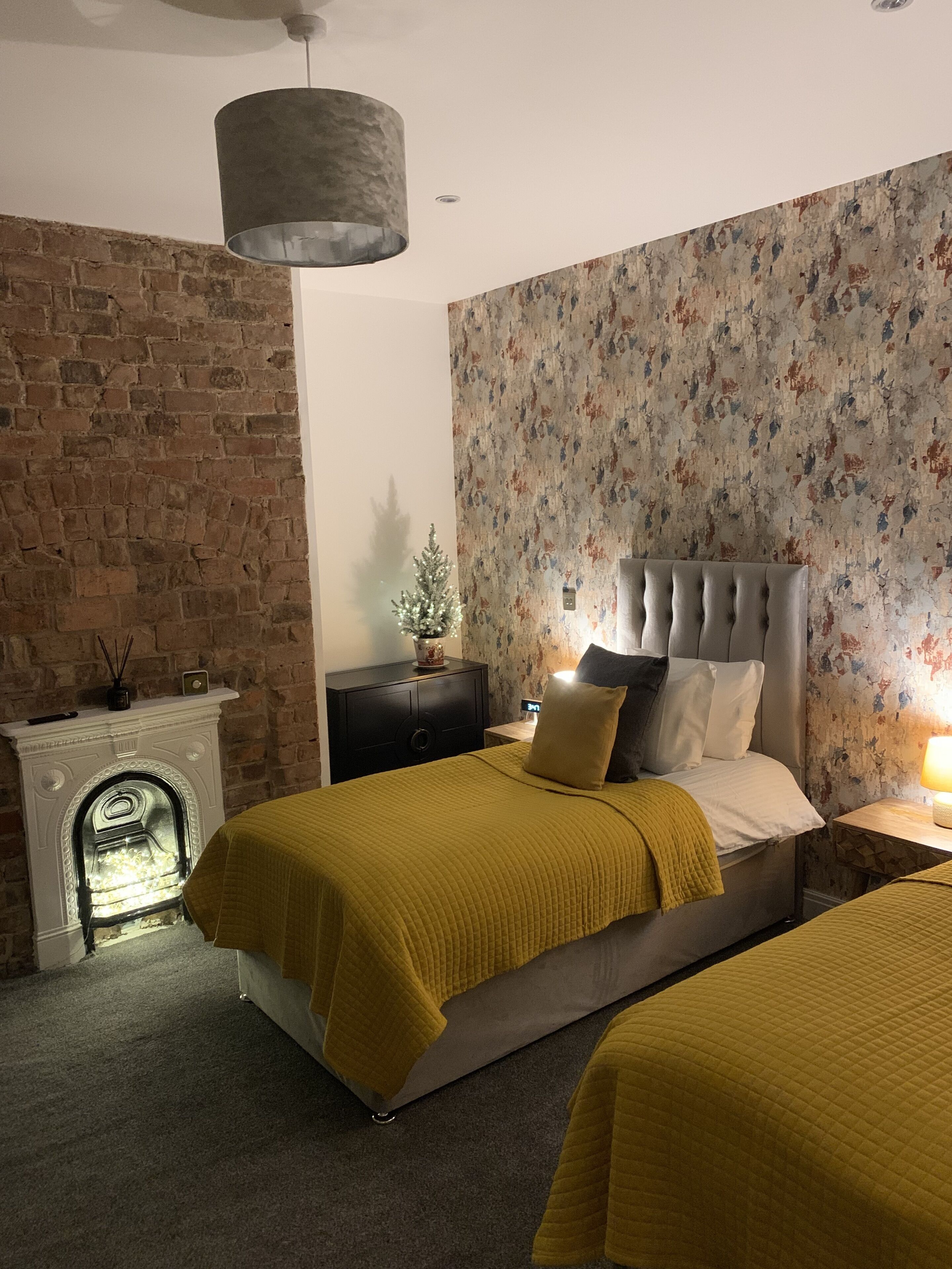Ein Schlafzimmer mit Ziegelwand, Kamin, einem Bett mit Quastensteppung und floralem Wandpapier.