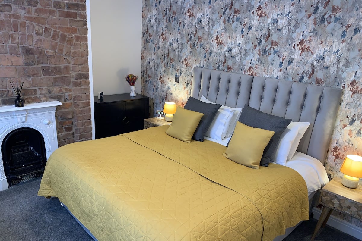 A bedroom with a large bed, a fireplace, a nightstand with a lamp, and a floral wallpaper.