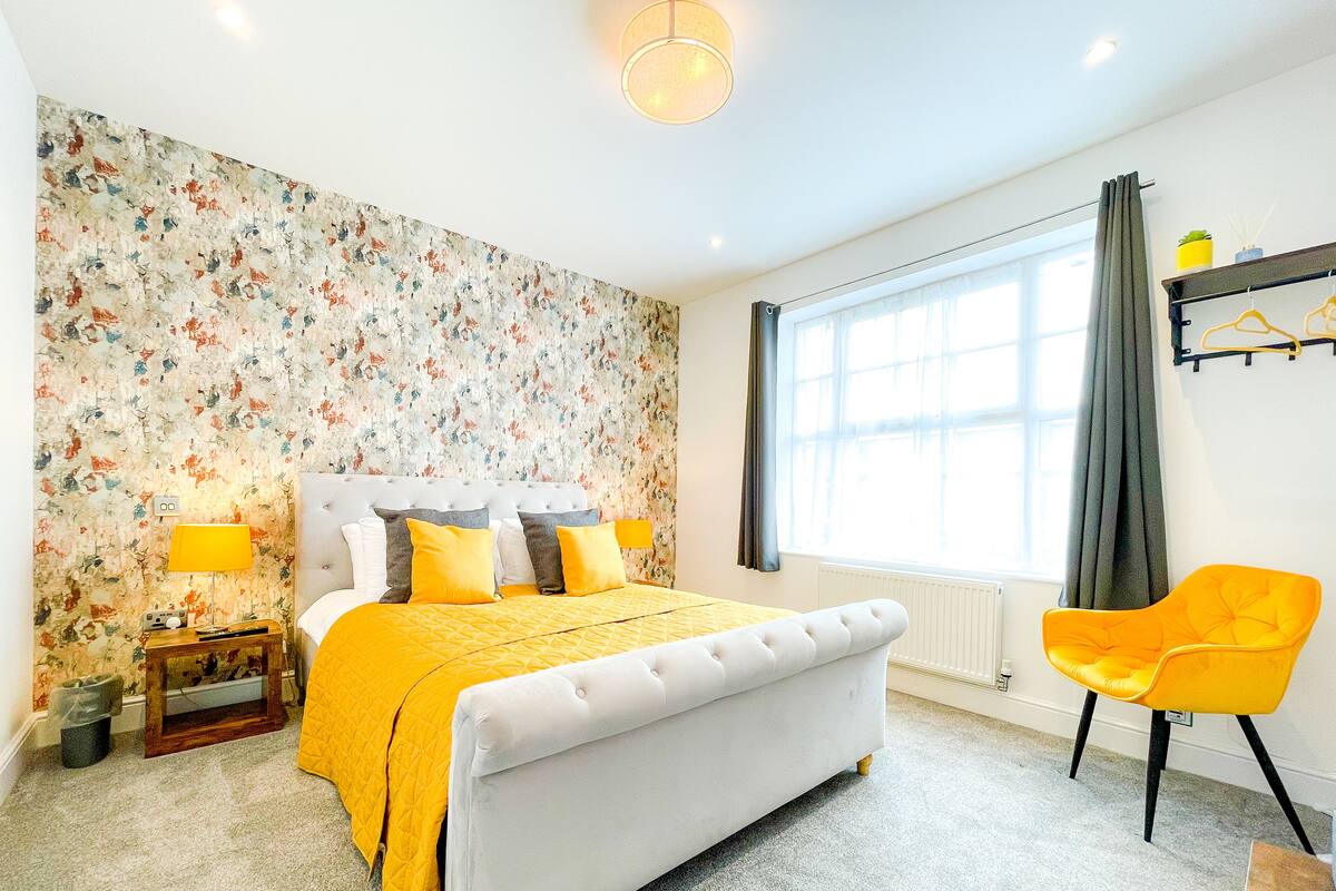 A bedroom with a bed, a bedside table, a lamp, a chair, and a window with curtains.