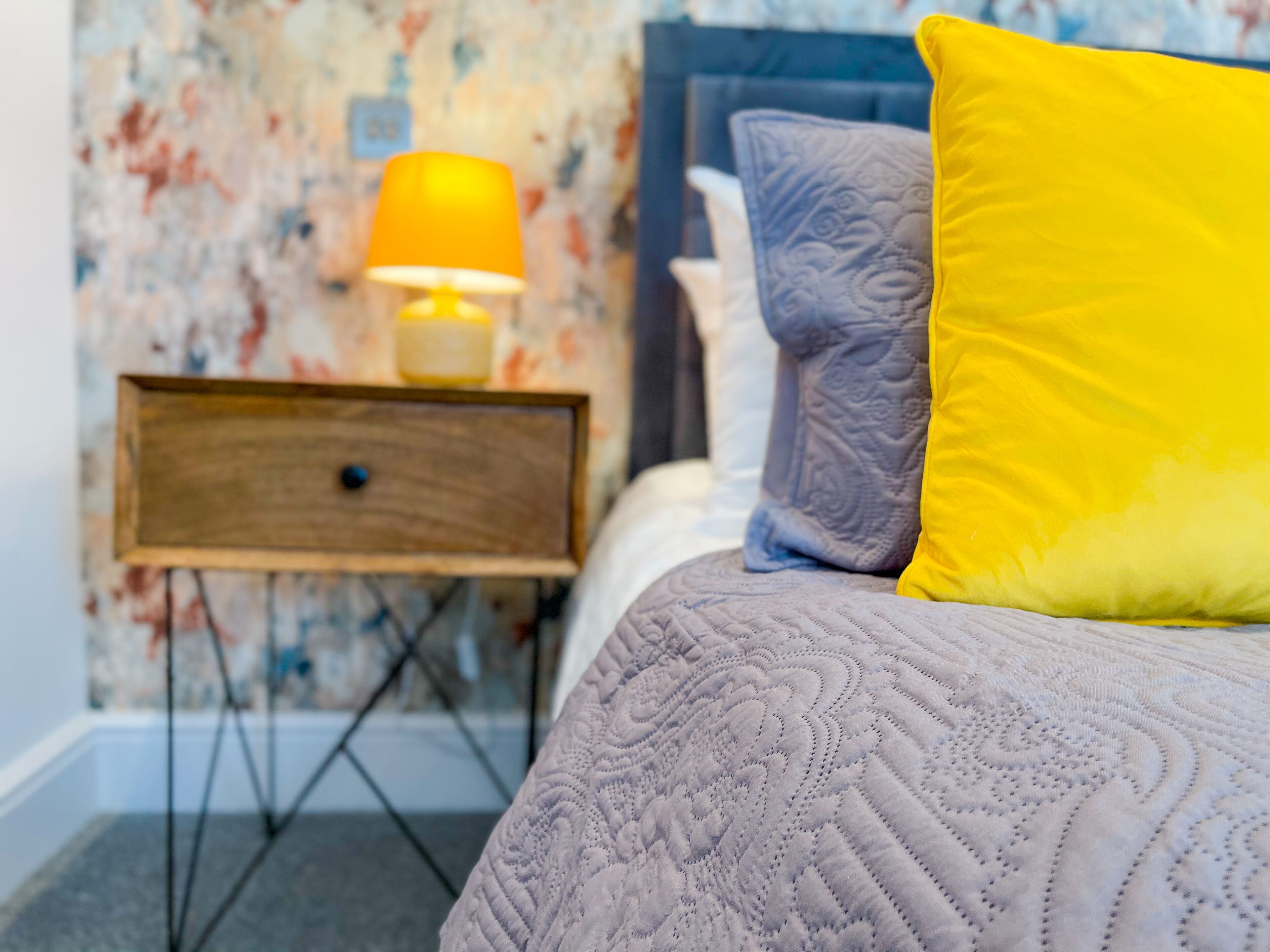 A bed with a patterned duvet, a yellow pillow, and a grey pillow. There is a nightstand with a lamp and a small drawer. The wall has a floral wallpaper.