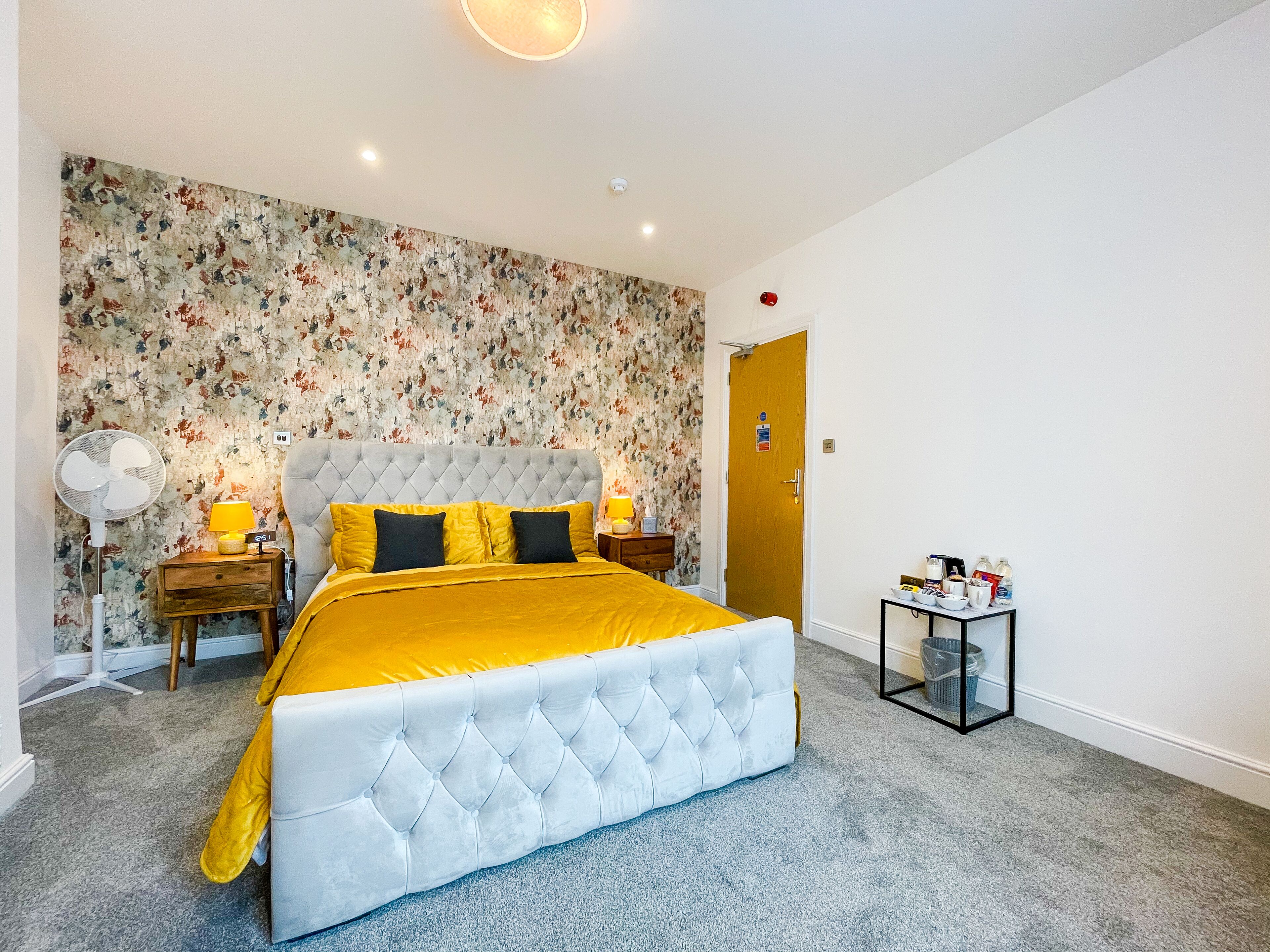 A bedroom with a tufted headboard, a bed with yellow bedding, a floral wallpaper, a bedside table with a lamp, and a small table with items.