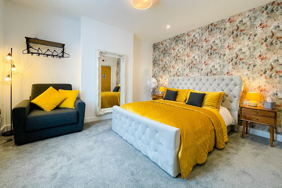 A bedroom with a large bed, a chair, a mirror, a nightstand, and a wall with floral wallpaper.