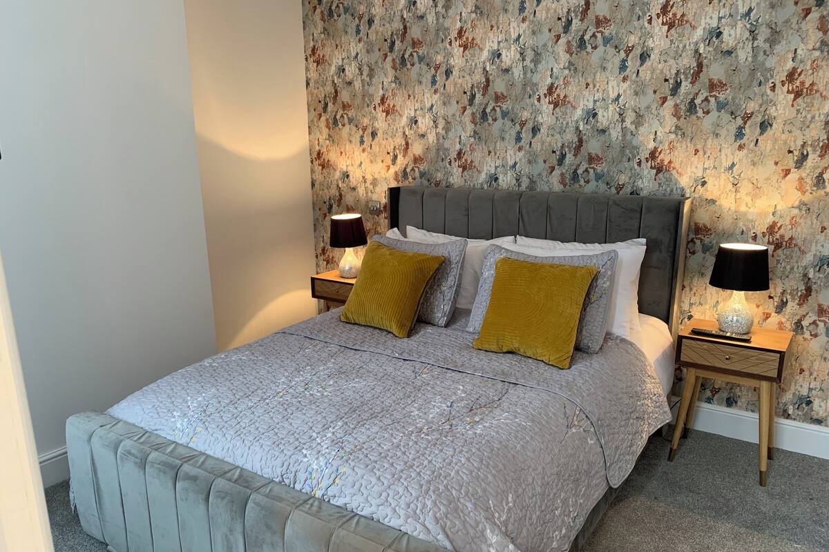A bedroom with a grey upholstered bed, two bedside lamps, a wooden nightstand, and a floral wallpaper.
